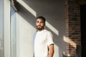 Bearded man in white t shirt looking at camera while standing near window in sunlit loft workplace