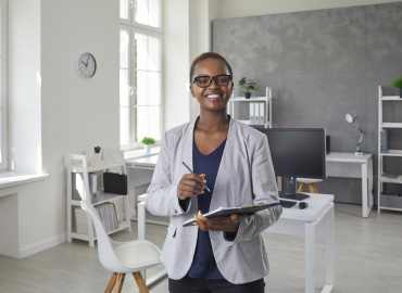 Portrait of happy successful satisfied businesswoman at work. Beautiful black woman in jacket with short hair looking at camera and smiling standing in office interior with computer desk in background